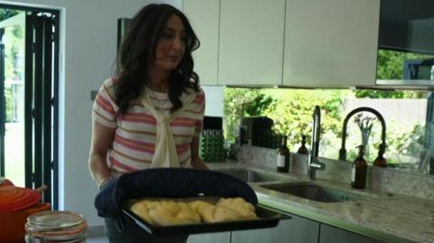 Joanna holds a baking tray containing fresh challah bread in a kitchen.