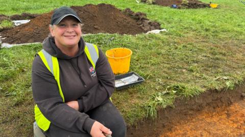 A woman sits in a field on the side of a dug out trench. She is dressed in black with a hi-vis vest on. behind her sits mounds of dug out soil and yellow buckets. She is smiling widely and holds a trowel in one hand.