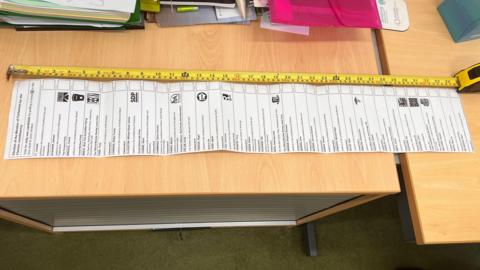 A long sheet of paper sits on wood-topped tables, with various boxes printed on it. A yellow tape measure is extended parallel to the paper.