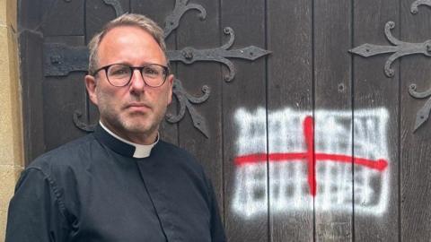 The Reverend Michael Smith looks serious as he stands in front of the large oak doors where a St George flag is roughly sprayed in red and white paint in the centre. He has short blond hair and round black-rimmed glasses and is wearing a black ecclesiastical shirt and white collar.