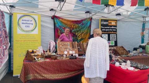 A market stall with three tables full of items for sale including jewellery and crystals. There are two banners in the back advertising Ahead of Wellbeing's services. A woman stands behind the stall in a purple t-shirt and brown necklace smiling as she serves a female customer with her back to the camera with blonde hair and wearing a long white cardigan.