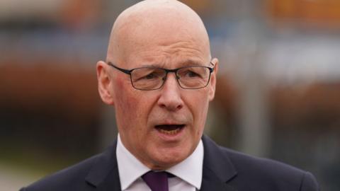 John Swinney, mid speech, wearing black spectacles, a black blazer, a white shirt and a purple tie. The shot captures him from the shoulders up.