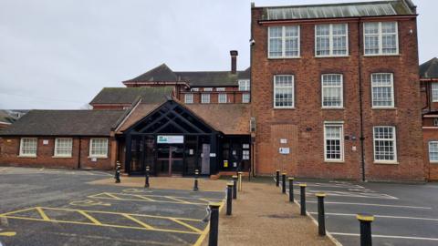 A brick building with three storeys on the right one storey on the left. There is a car park and a path in front.