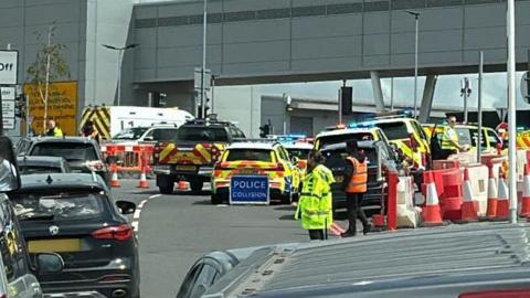 A queue of cars runs to the side of a police sign and emergency vehicles and workers.