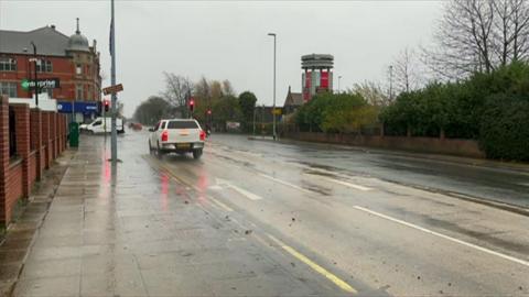 Cars are travelling along a road in the rain, with traffic lights on red and some buildings either side of the road.