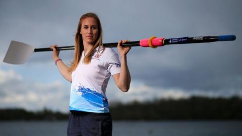 Helen Glover, standing outdoors near a body of water, holding a rowing oar across her shoulders. She has long, light brown hair and is dressed in sportswear, including a white top with blue accents and branding. The background is an overcast sky with distant trees.