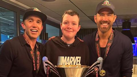 Aiden Waller stands in front of a large national league trophy in between Ryan Reynolds (right) and Rob McElhenney (left). All three of them smile at the camera. Both Ryan and Rob wear Wrexham black caps and dark long sleeve t-shirts, while Aiden, who has short dark hair, wears a black Marvel hoodie. 