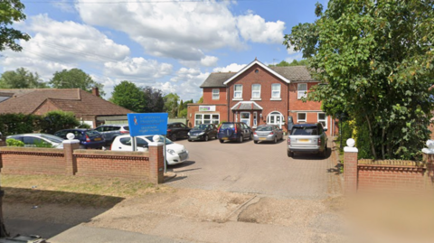 A google maps view of detached building that has about a dozen parking spaces and low brick walls. A blue sign is at the entrance