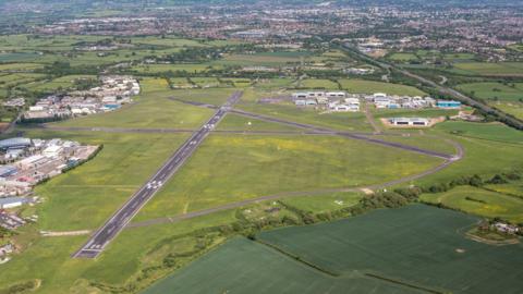 Aerial image of Staverton Airport - a small airport - in Gloucestershire. There are three runways that intersect one another on an airfield with hangars to the right. Cheltenham can be seen behind.