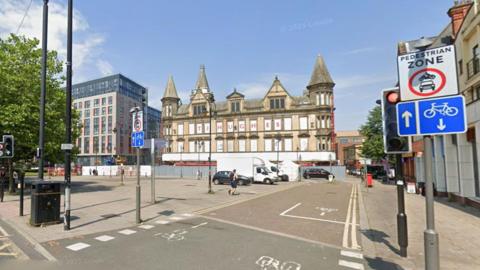A large paved area with trees on one side and a boarded up yellow brick building which has a sign in red lettering which reads TJ Hughes. A white road sign with black writing which reads pedestrian zone and a blue road sign showing a cycle lane and an image of a bicycle and arrows are next to a set of traffic lights.