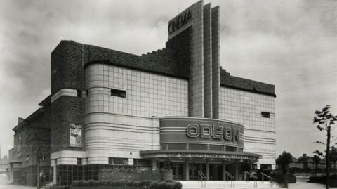 An Odeon building which has an Art Deco structure with rounded corners and horizontal lines. There is a central tower rising above the entrance with horizontol straight lines. At the bottom “ODEON” is displayed. There are steps leading up to the doors. 