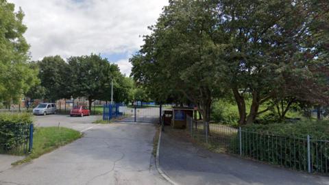 An exterior view of the entrance to Mill Rythe Infant School, there are primary blue coloured gates on a road, with a car park to the left which has a red fiat parked and a silver people carrier, there are trees lining either side of the road