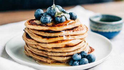 A stack of pancakes and blueberries being drizzled with maple syrup. 