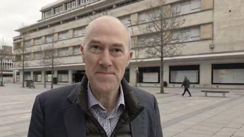 A man called Harry Holt stands outside a former department store in Plymouth city centre. The building is large and appears to be made from sandstone blocks. Holt is a bald man wearing a black suit jacket over a dark Barbour jacket and collared shirt.