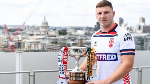 George Williams stands in front of the London skyline with the Ashes trophy wearing full England rugby league strip