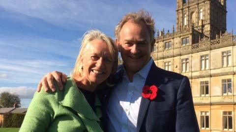 Sally Taylor and Hugh Bonneville stood outside of a large manor house.