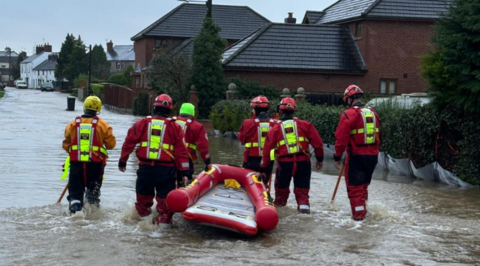 A red boat being dragged through a flooded street by six people in red coats and trousers