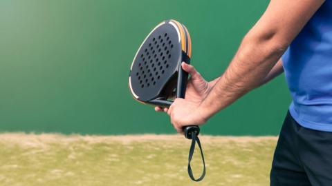 Someone holding a black padel racket which has holes in the middle of it and orange around the sides. They are holding it with both hands and wearing a blue t shirt. You can't see their face but behind is green tarmac and a darker green wall.