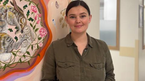 Jenna Stuchfield wears a khaki coloured shirt and has dark hair tied back. She is stood in the maternity ward next to her mural.