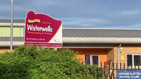 The front of Waterwells Primary Academy - a new build primary school. There is a bush and a metal fence at the front and a large red sign with the name of the school on it. The walls are brick and rendered orange.
