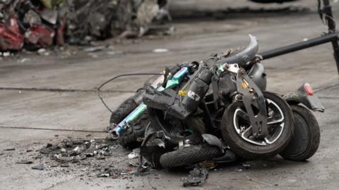 A pile of crumpled up e-scooters and e-bikes. They are all black in colour apart from one which is a two toned green and purple colour. Behind them is a section of a much larger pile of crushed metal.