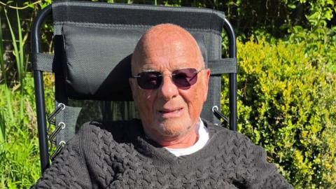 An older man wearing sunglasses and a grey jumper which is pulled over a white T-shirt. He is sitting in a chair outside with bushes behind him. 