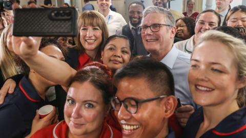 Close up Sir Keir Starmer and Rachel Reeves posing for a selfie taken by an NHS staff member. They are smiling and surrounded by a group of NHS staff all smiling at the camera