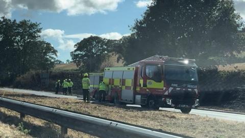 Firefighters and a fire engine are at the side of the road and charred vegetation can be seen.
