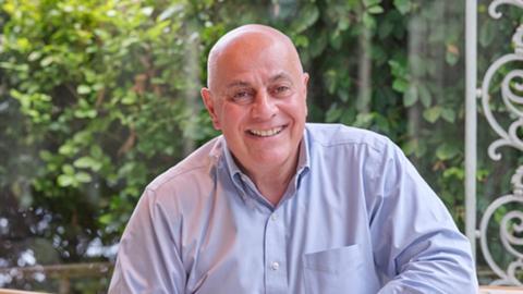 White man sitting inside a restaurant conservatory. Blue shirt, smiling at the camera, bald head, brown eyes and prominent eyebrows. 