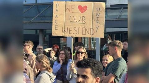 A woman holds a sign saying 'Peace, Unity, We love our West End'