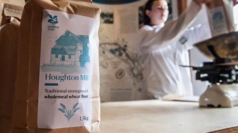 A bag of flour is sitting on a surface. The bag is brown and there is a label on it showing an image of a mill, the National Trust emblem and the name of the product. In the blurred background a woman can be seen measuring flour on an old-fashioned set of scales.