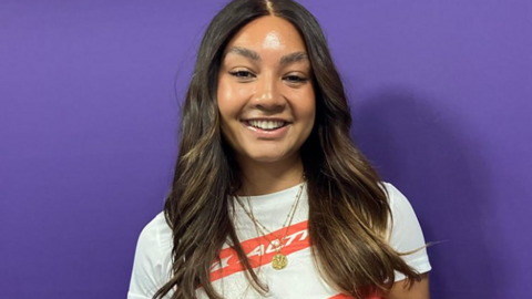 Lillian Istance pictured in front of a purple background. She has long brown hair and is wearing a white and red t-shirt with a gold necklace.