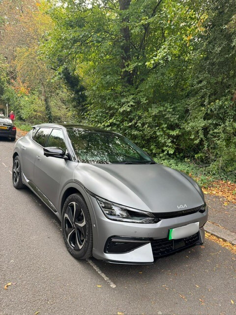 A grey Kia EV6 coupe style car is parked in a parking bay on a road. Trees and a footpath can be seen behind the car.