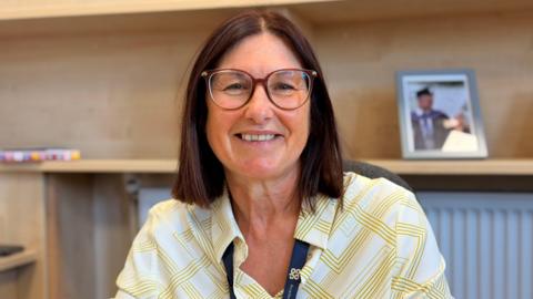 A women with short brown hair, glasses and a yellow and white patterned shirt. She is looking at the camera smiling.
