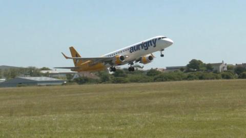 A yellow and white airplane with Aurigny logo on its side, setting off into the sky from the take off area at the airport. Blue skies and green grass visible.