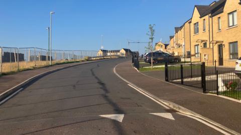 A line of houses which are light coloured. There is a metal fence on the other  side of the road and a shrubby area. 