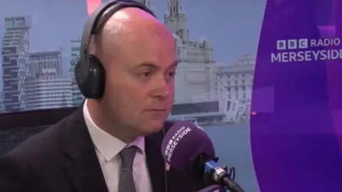 Liam Robinson, wearing black headphones and a black suit, white shirt and grey tie, sits in front of a purple microphone in a BBC Radio Merseyside studio