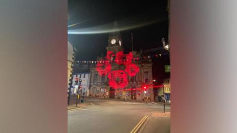 Kendal Town Hall is an impressive stone building with a central clock tower, arch windows on the ground floor and high rectangular ones on the first floor. It sits on a road junction and is attached to an old white fronted building. It is shown at night with a circle of five poppies beamed on to its front.