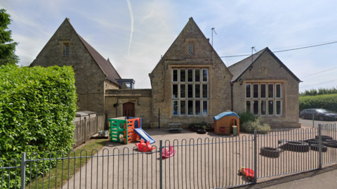 Stone school buildings with steep gabled roofs surround a fenced outdoor play area containing small plastic slides and toys.
