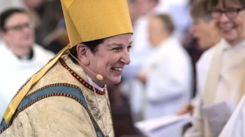 Bishop Anne Dyer dressed in full religious wear, and wearing a small head microphone, smiling while shaking hands with someone. Other people are gathered in background.
