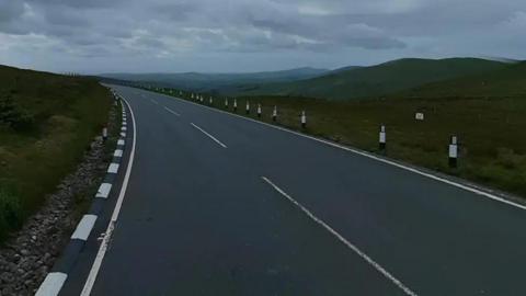 The Mountain Road just beyond the Brandywell Junction, which has a grass embankment to the left, and hills to the right. 