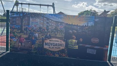 Banner on a play park fence, showing children playing sunshine and men being rejected at a barrier. 