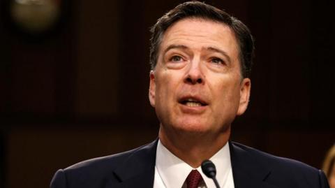 Former FBI Director James Comey, wearing a blue suit over a white shirt and red ties, testifies before a Senate Intelligence Committee in 2019