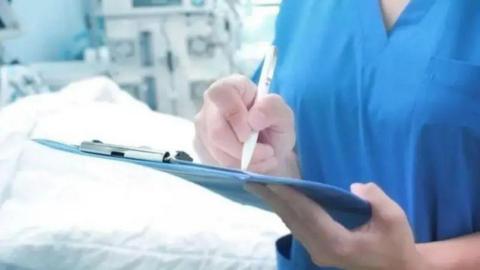 A close-up of a nurse wearing blue scrubs in a hospital room, she is writing on a blue clipboard and there is a bed in the background.