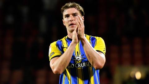 James Bree claps fans with hands to side of his face, wearing blue and yellow away kit