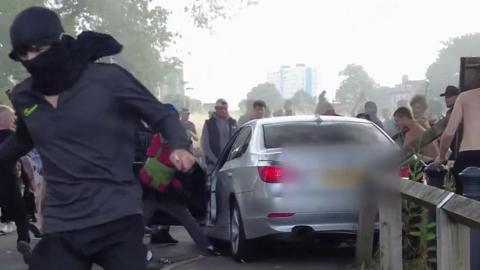 Man wearing a mask running from a silver BMW, which is being attacked by a group of white men, two of whom on the right are topless