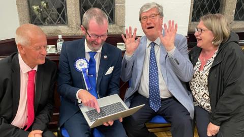 Four people - three men and a woman - wearing smart clothing sit on chairs to look at a laptop. One of the men is holding his hands up.