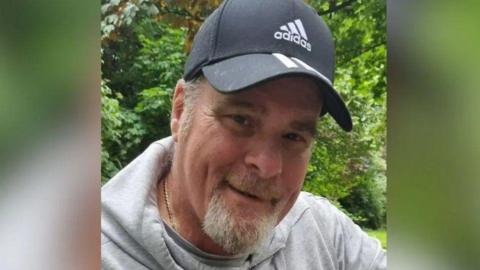 A man in his sixties with a goatee and a baseball cap. He is wearing a grey hoodie. 