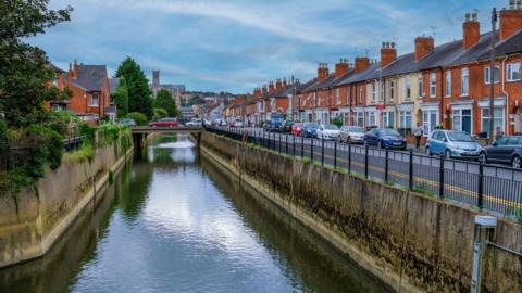 Looking along the Sincil Dyke waterway and neighbouring road towards Lincoln Cathedral. To the right is a line of terraced red-brick houses, some painted in shades of cream, with cars parked outside 