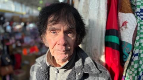 Paul is stood at his stall with clothing hanging in the background.He has a grey jacket on over a light-coloured fleece. He has dyed dark hair which is greying near his face.